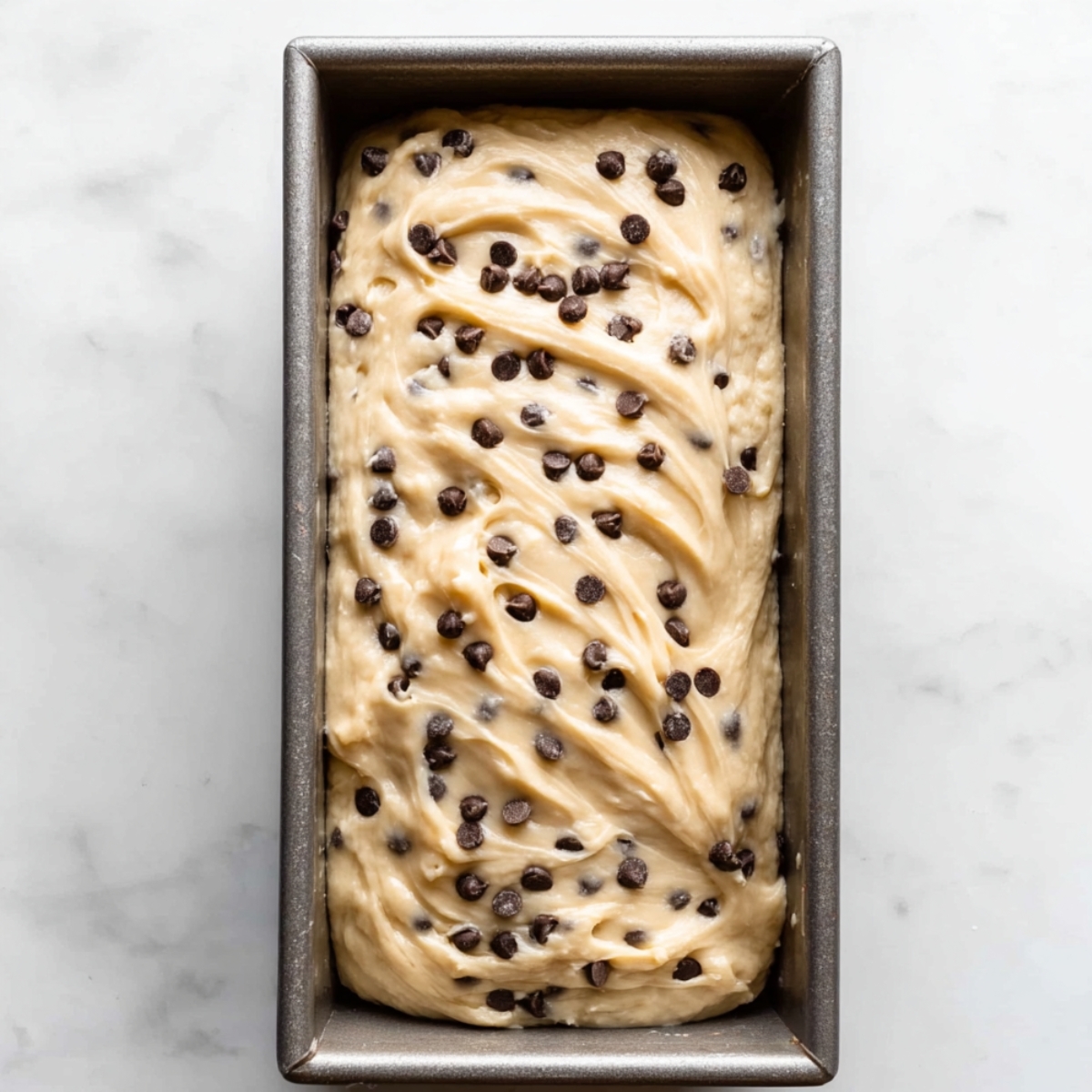 Chocolate Chip Loaf Cake Recipe 11 Chocolate chip loaf cake batter spread into a metal loaf pan, topped with extra mini chocolate chips and swirled smooth before baking.