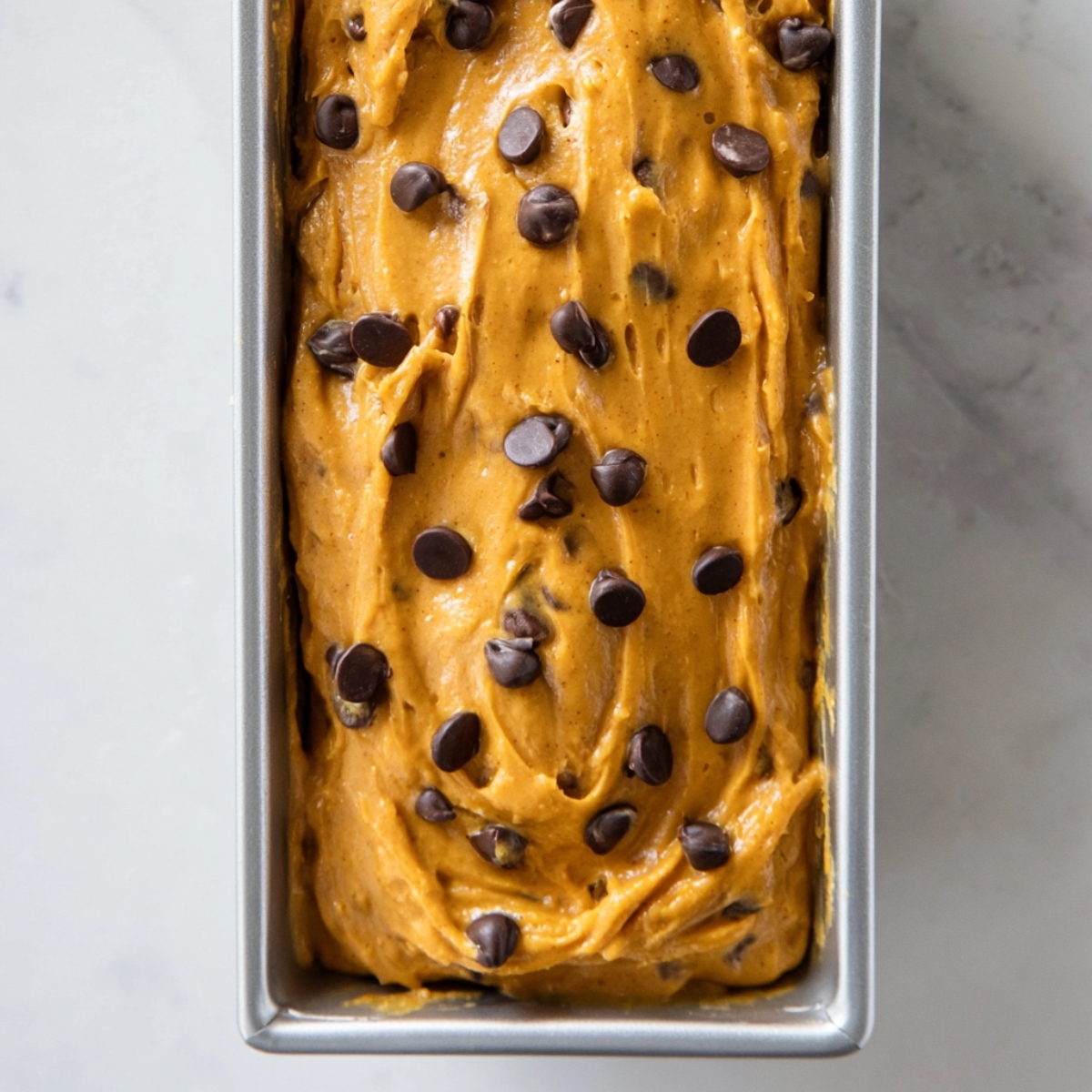 Pumpkin Chocolate Chip Bread Recipe 11 Pumpkin chocolate chip bread batter spread in a loaf pan, topped with chocolate chips and ready for baking on a light marble surface.
