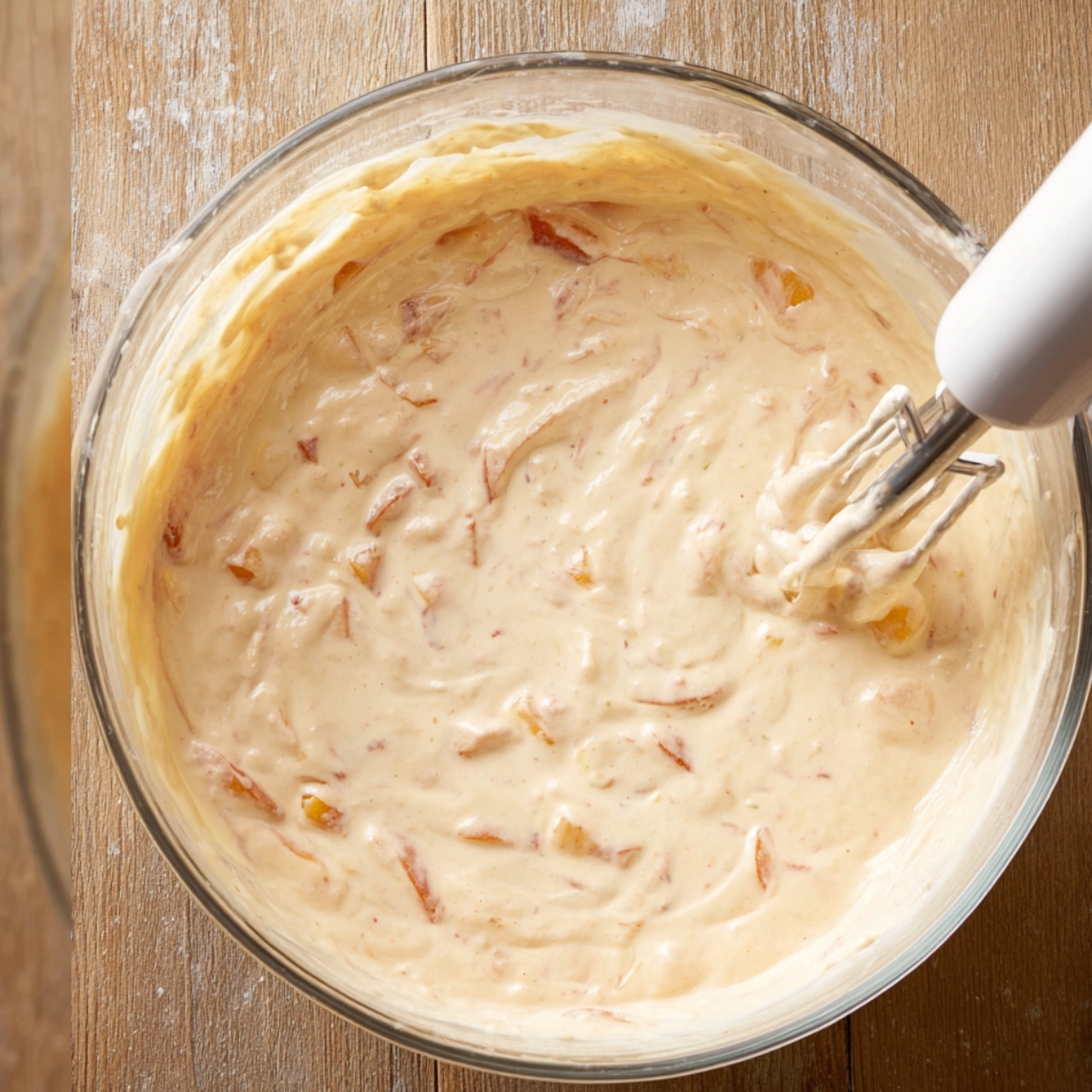 The Best Peach Cheesecake Recipe 11 Creamy peach cheesecake filling mixed in a glass bowl with visible peach pieces and a hand mixer, ready to be poured into a crust.