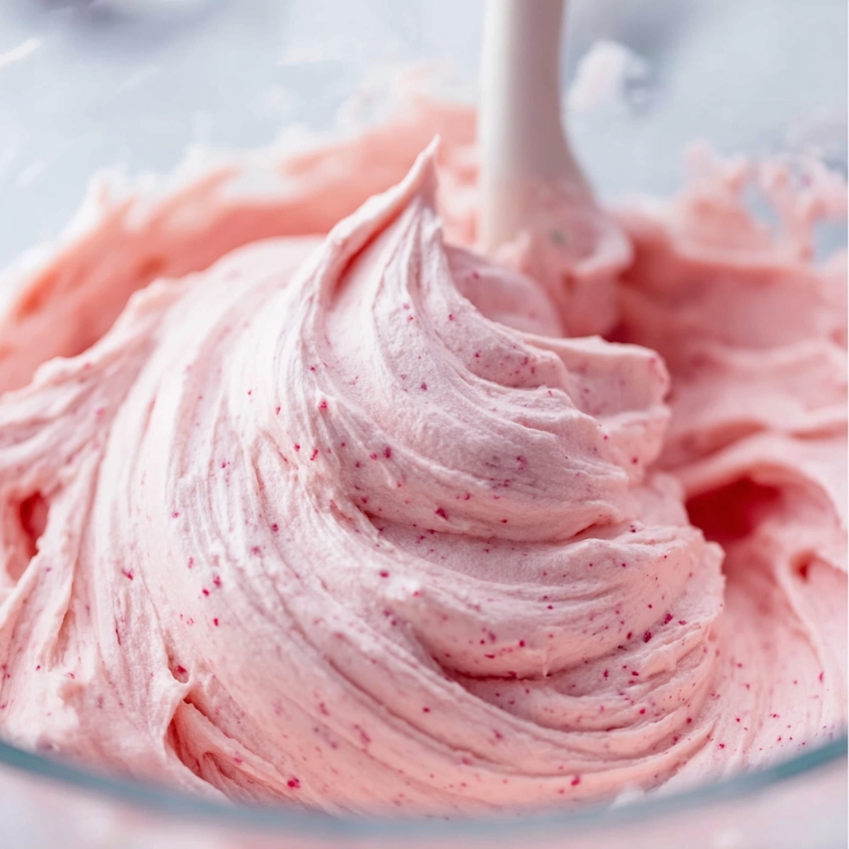 Strawberry Nutella Cupcakes Recipe 11 Smooth strawberry buttercream frosting with pink berry speckles being whipped in a glass mixing bowl, showing fluffy swirls and creamy texture.