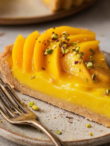 Easy Mango Curd Tart Recipe 1 Slice of Mango Curd Tart topped with fresh mango slices and chopped pistachios on a rustic ceramic plate, featuring a buttery crust, creamy golden filling, and vintage dessert fork.