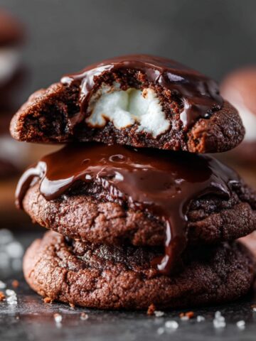 Marshmallow-Surprise Hot Cocoa Cookies 2 Stack of fudgy Marshmallow-Surprise Hot Cocoa Cookies topped with glossy chocolate glaze, featuring a gooey marshmallow center and scattered cookie crumbs on a dark surface.