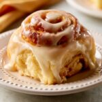 Freshly baked Orange Sweet Roll topped with creamy glaze, served on a white decorative plate, showcasing fluffy layers and rich icing dripping over the sides.