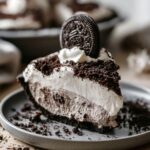 Slice of Oreo Pudding Pie topped with whipped cream, Oreo cookie crumbs, and a whole Oreo cookie garnish on a gray dessert plate, with a chocolate cookie crust and creamy cookies-and-cream filling.