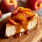 Slice of peach cheesecake topped with glossy caramelized peach slices and rich peach sauce, served on a wooden board with fresh peaches in the background.