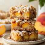 Stack of homemade Peach Pie Crumb Bars topped with buttery streusel and vanilla glaze on a ceramic plate, with fresh peaches in the background.