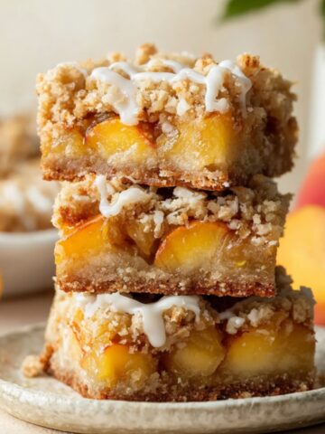 Easy Peach Pie Crumb Bars Recipe 1 Stack of homemade Peach Pie Crumb Bars topped with buttery streusel and vanilla glaze on a ceramic plate, with fresh peaches in the background.