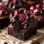 Stacked fudgy Roasted Chocolate Cherry Brownies topped with glossy roasted cherries and melted chocolate chips, featuring a rich gooey center and cherry compote dripping over the sides on parchment paper.