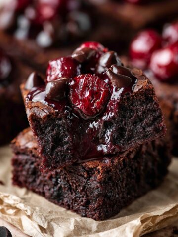 Roasted Chocolate Cherry Brownies 1 Stacked fudgy Roasted Chocolate Cherry Brownies topped with glossy roasted cherries and melted chocolate chips, featuring a rich gooey center and cherry compote dripping over the sides on parchment paper.