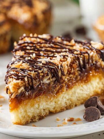 Slice of Samoa Cookie Cake topped with toasted coconut caramel and chocolate drizzle, featuring a soft cookie base and gooey caramel layer.