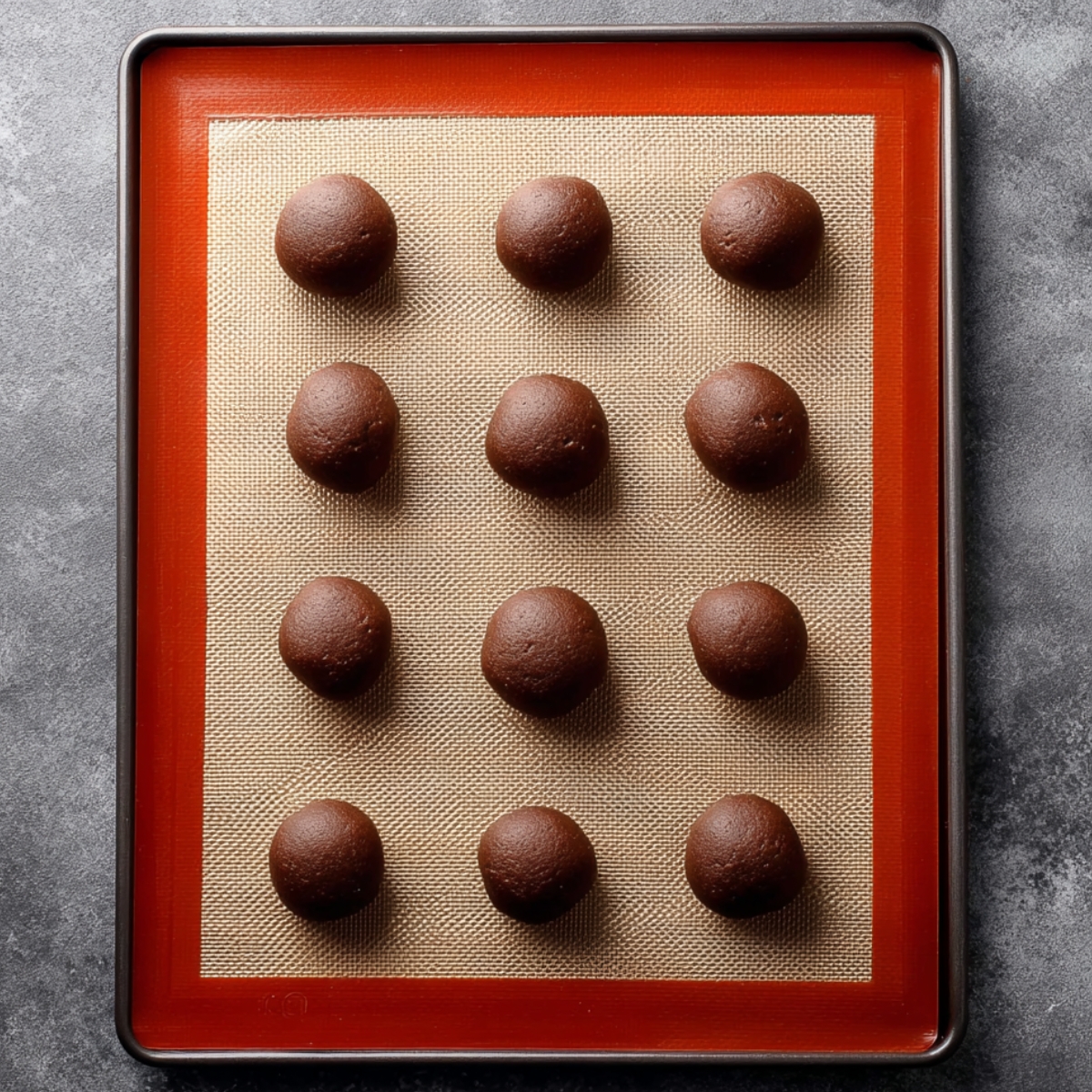 Marshmallow-Surprise Hot Cocoa Cookies 11 Chocolate cookie dough balls evenly spaced on a silicone-lined baking sheet, ready for baking, photographed from above on a gray stone countertop.