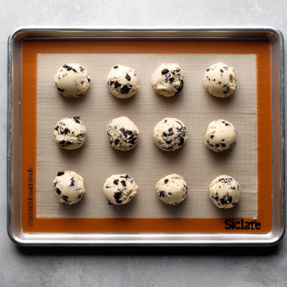 Easy Cookies and Cream Cookies 11 Cookies and cream cookie dough balls arranged in neat rows on a silicone-lined baking sheet, ready for baking, photographed from above on a light gray surface.