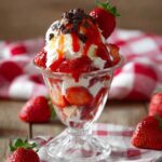 Strawberry Sundaes served in a vintage glass dessert cup with layers of vanilla ice cream, whipped cream, fresh strawberries, strawberry sauce, and chocolate shavings on a rustic table.