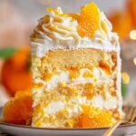Tall slice of Sunshine Citrus Cake filled with whipped cream and mandarin oranges, topped with piped frosting and fresh orange segments on a dessert plate.