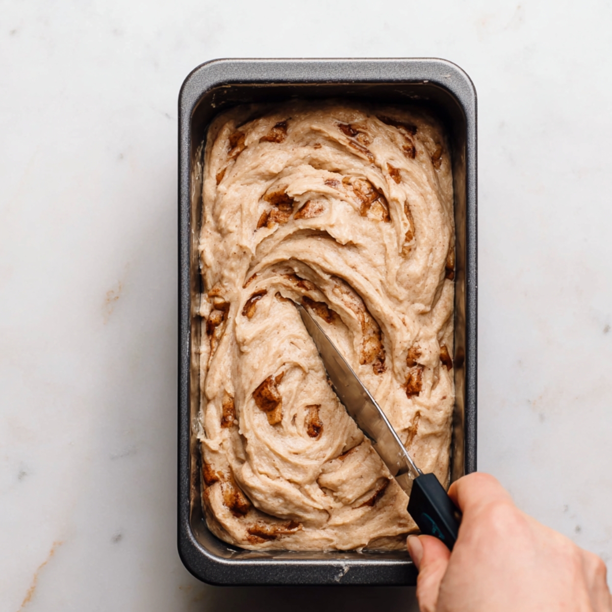 Apple Cinnamon Quick Bread Recipe 11 Overhead view of apple cinnamon bread batter swirled with a knife in a metal loaf pan, creating marbled cinnamon patterns before baking.