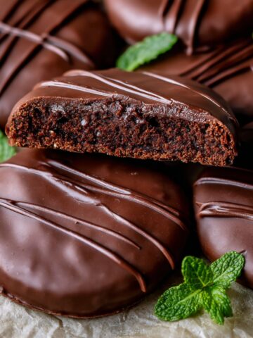 Homemade Thin Mint Cookies Recipe 1 Close-up of homemade Thin Mint Cookies coated in glossy dark chocolate, one cookie sliced in half to reveal the crisp chocolate cookie center, garnished with fresh mint leaves.