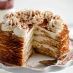 Slice-cut Tiramisu Crepe Cake showcasing dozens of delicate crepe layers filled with mascarpone cream, topped with whipped cream rosettes and cocoa powder on a white cake stand.