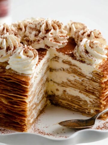 The Best Tiramisu Crepe Cake Recipe 1 Slice-cut Tiramisu Crepe Cake showcasing dozens of delicate crepe layers filled with mascarpone cream, topped with whipped cream rosettes and cocoa powder on a white cake stand.
