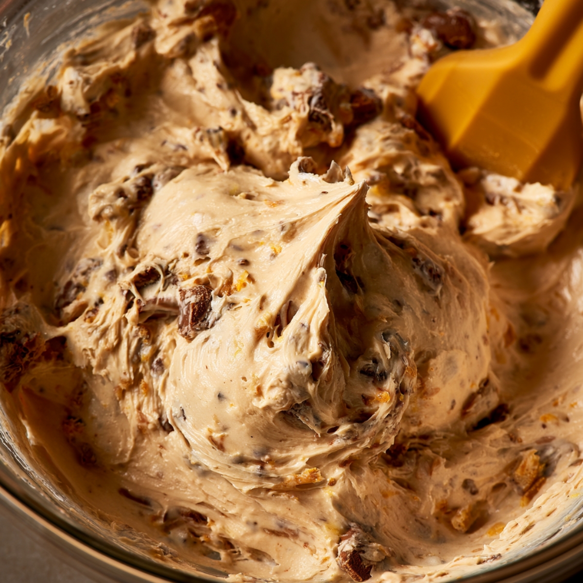 Easy Peanut Butter Butterfinger Pie Recipe 10 Thick peanut butter cheesecake filling being folded with chopped Butterfinger candy pieces in a glass mixing bowl using a yellow spatula.
