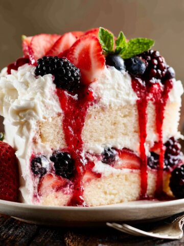Easy Berries and Cream Cake Recipe 1 Slice of Berries and Cream Cake layered with fresh strawberries, blackberries, blueberries, whipped cream frosting, and berry sauce on a dessert plate.
