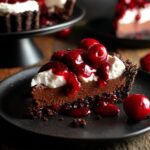 Slice of Black Forest Tart topped with whipped cream and cherry compote on a black dessert plate