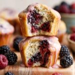 Stacked Blackberry Lemon Poppy Seed Muffins with pink lemon glaze and juicy berry-filled centers on a rustic wooden board.