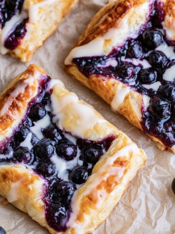 The Best Blueberry Danish Recipe 2 Freshly baked Blueberry Danish pastries with flaky puff pastry, cream cheese filling, homemade blueberry topping, and vanilla glaze on parchment paper.
