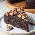 Slice of rich Chocolate Hazelnut Cake topped with roasted hazelnuts and dark chocolate chunks.