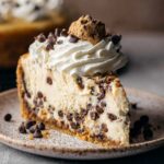 Creamy cookie dough cheesecake slice topped with whipped cream, mini chocolate chips, and a mini chocolate chip cookie.