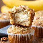Cream cheese banana muffins with a creamy cheesecake center and crunchy pecan topping, one muffin cut open to reveal the filling.