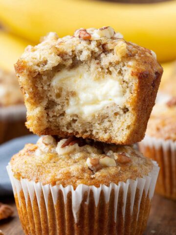 Easy Cream Cheese Banana Muffins 2 Cream cheese banana muffins with a creamy cheesecake center and crunchy pecan topping, one muffin cut open to reveal the filling.