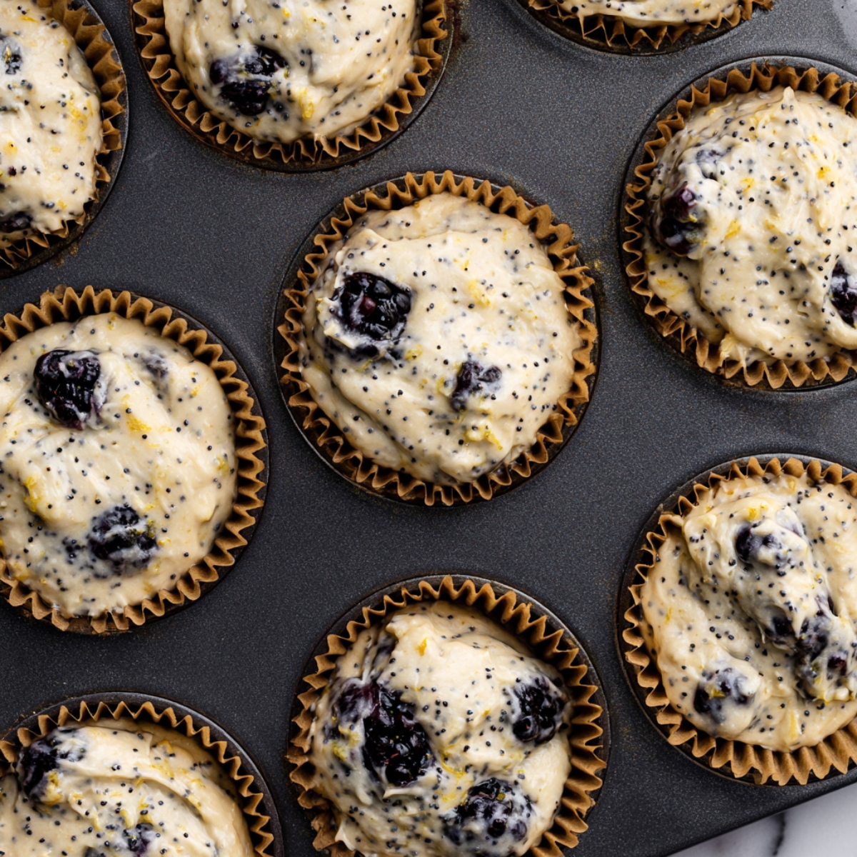 Blackberry Lemon Poppy Seed Muffins 11 Overhead view of blackberry lemon poppy seed muffin batter portioned into brown paper liners in a dark muffin pan before baking.