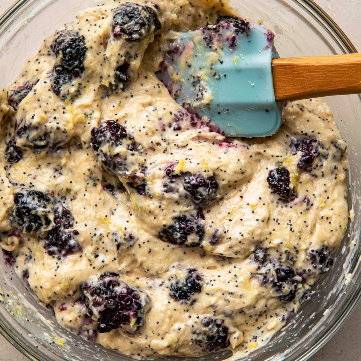 Blackberry Lemon Poppy Seed Muffins 10 Close-up of thick blackberry lemon poppy seed muffin batter being folded with a blue silicone spatula in a glass mixing bowl.