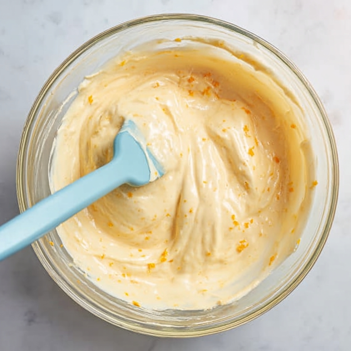 Easy Orange Mousse Cups Recipe 11 Folding whipped egg whites into creamy orange mousse mixture with a blue silicone spatula in a clear glass mixing bowl.