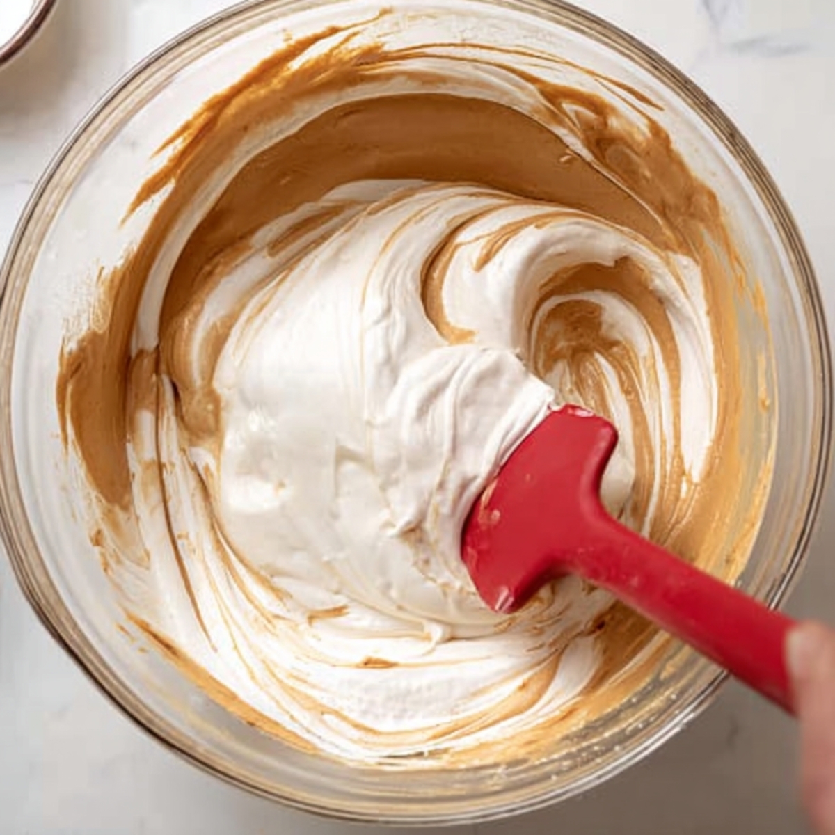 Easy Speculoos Tiramisu Recipe 11 Folding whipped cream into Biscoff mascarpone mixture with a red silicone spatula in a glass mixing bowl.