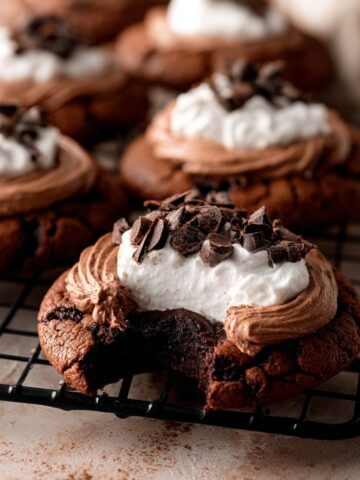 Easy French Silk Pie Cookies Recipe 2 Homemade French Silk Pie Cookies topped with chocolate mousse, whipped cream, and chocolate curls on a cooling rack, with one cookie showing a fudgy bite.