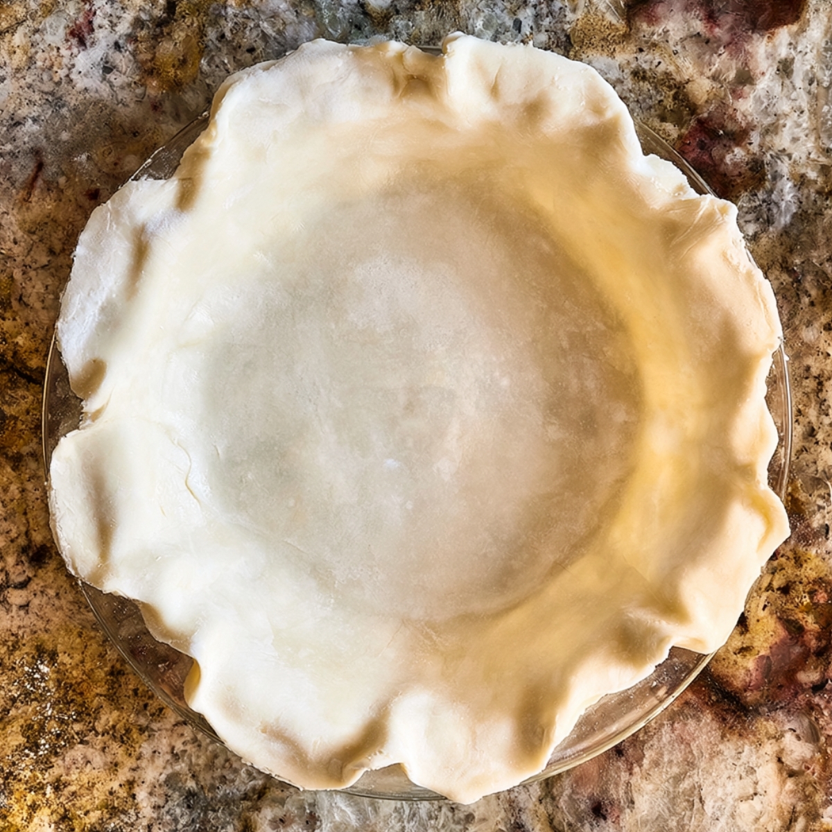 Easy Huckleberry Pie Recipe 10 Top-down view of an unbaked pie crust pressed into a glass pie pan on a granite countertop, ready for huckleberry pie filling.
