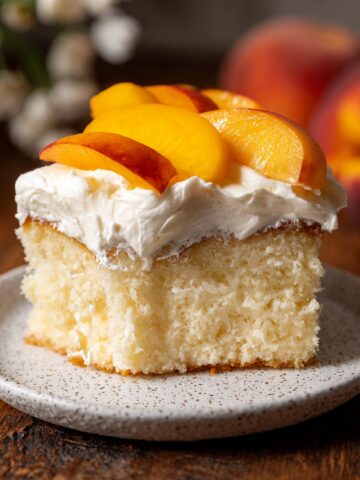 Best Peaches and Cream Cake Recipe 1 Slice of Peaches and Cream Cake topped with whipped cream and fresh peach slices on a speckled ceramic plate.