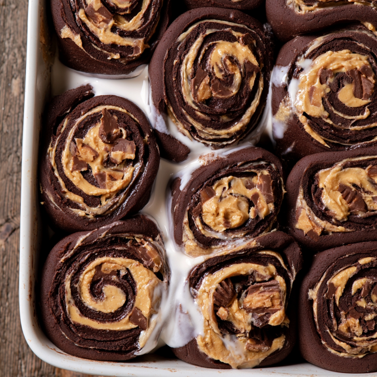 Chocolate Peanut Butter Rolls Recipe 11 Chocolate peanut butter rolls in a baking dish with rich chocolate dough, creamy peanut butter filling, chopped peanut butter cups, and sweet icing between the rolls.