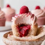 Bitten Raspberry Filled Cupcakes topped with raspberry cream cheese frosting, fresh raspberry, and freeze-dried raspberry powder, revealing a gooey raspberry jam center.