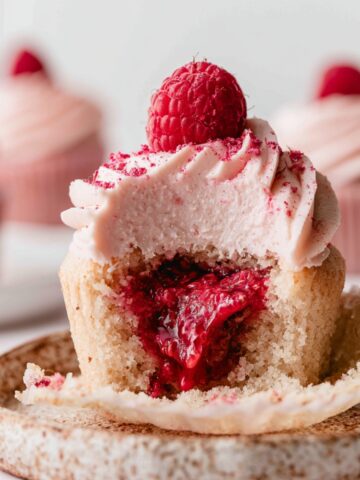 Best Raspberry Filled Cupcakes Recipe 2 Bitten Raspberry Filled Cupcakes topped with raspberry cream cheese frosting, fresh raspberry, and freeze-dried raspberry powder, revealing a gooey raspberry jam center.