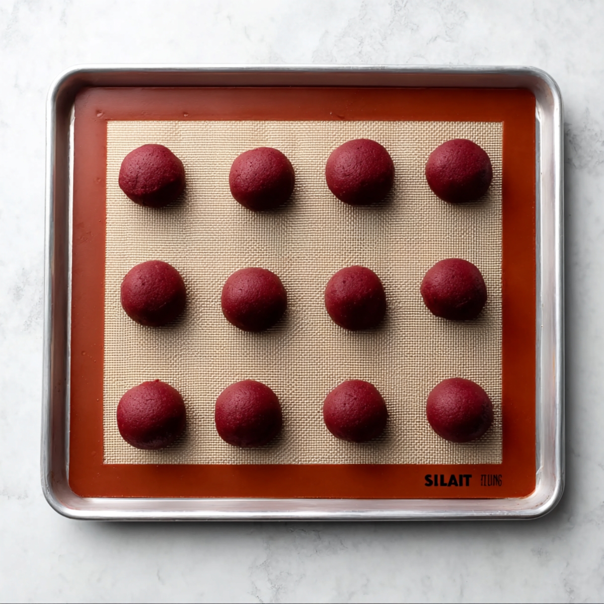 White Chocolate Red Velvet Cookies Recipe 11 Red velvet cookie dough balls arranged on a silicone baking mat on a baking sheet, ready to bake.