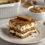 Slice of homemade Speculoos Tiramisu topped with crushed Biscoff cookies and a whole biscuit on a white dessert plate.