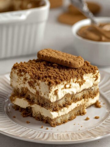 Easy Speculoos Tiramisu Recipe 2 Slice of homemade Speculoos Tiramisu topped with crushed Biscoff cookies and a whole biscuit on a white dessert plate.