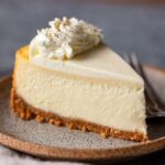 Slice of Vanilla Bean Cheesecake with a buttery graham cracker crust and whipped cream topping on a ceramic dessert plate.