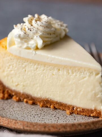 Easy Vanilla Bean Cheesecake Recipe 1 Slice of Vanilla Bean Cheesecake with a buttery graham cracker crust and whipped cream topping on a ceramic dessert plate.