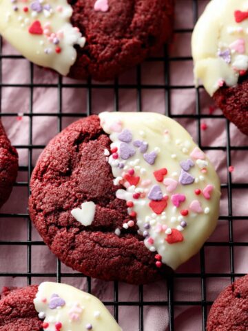 White Chocolate Red Velvet Cookies Recipe 2 Soft White Chocolate Red Velvet Cookies and decorated with Valentine's Day heart sprinkles on a cooling rack.