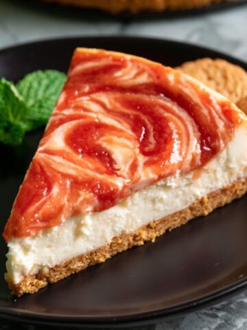 The Best Guava Cheesecake Recipe 1 Slice of creamy guava cheesecake with red guava swirl topping on a black plate with mint garnish and cookies