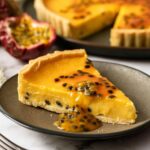 Slice of homemade Passionfruit Tart with silky passion fruit custard and glossy passion fruit topping served on a dark ceramic plate with the whole tart in the background.