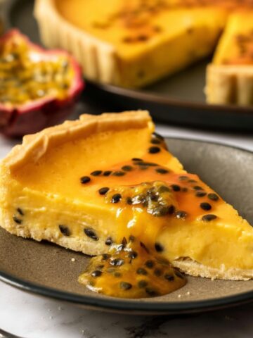 The Best Passionfruit Tart Recipe 4 Slice of homemade Passionfruit Tart with silky passion fruit custard and glossy passion fruit topping served on a dark ceramic plate with the whole tart in the background.